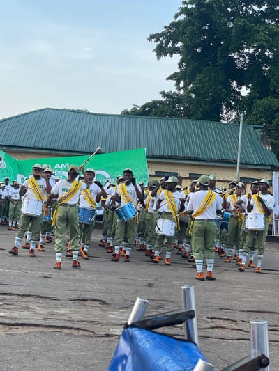 This media file depicts a youth band marching in a parade. The subjects are primarily young people, likely members of a youth corps, dressed in matching uniforms. They wear olive green pants, white shirts with yellow sashes, and green caps. Many are playing musical instruments, including drums (both white and blue), and brass instruments like trumpets. The band is marching on an asphalt surface, with a green corrugated metal roof building visible in the background. Some individuals are holding drumsticks and mallets, indicating they are actively playing.
The scene appears to be outdoors, likely during the daytime, given the natural light. The sky is visible, appearing mostly clear and bright, suggesting fair weather. The overall mood seems to be one of organized activity and performance, possibly a ceremonial event or a cultural celebration.
Visible text includes "AM" and "NYSC" on some of the uniforms and banners. The "NYSC" is likely an acronym for the National Youth Service Corps, a program in Nigeria that engages young graduates in national service. The context of Alagba, Nigeria, further supports this identification. The presence of a marching band and uniforms suggests a formal or semi-formal event. The individuals appear focused and engaged in their performance.