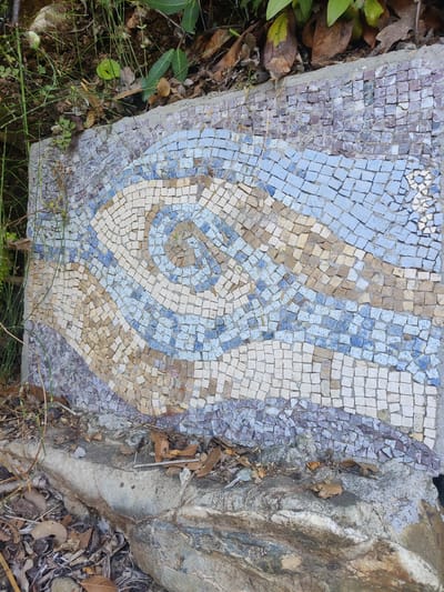 The image shows an outdoor mosaic on a flat surface, possibly a bench or a wall, in Δημοτική Ενότητα Ευρυμενών, Greece. The mosaic features a swirling pattern composed of small, square tiles in shades of blue, white, tan, and purple. The mosaic is partially obscured by vegetation, including grass and fallen leaves, and is situated on a rough, rocky surface. There are no people visible in the image.
