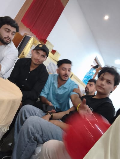 In the city of Khaniara, India, a group of five young men are gathered indoors, seated on the floor. The man on the far left wears a white shirt and has a beard. To his right, a man in a black shirt and a black baseball cap is visible. In the center, a man in a light blue shirt is speaking, with a red balloon obscuring parts of his body and the man to his right. The man to the right of the speaker wears a black t-shirt and has a yellow bracelet on his left wrist. The fifth man, partially obscured, is seated behind the man in the black t-shirt. The background features a red draped cloth and decorative golden paneling. The overall lighting suggests an indoor environment.