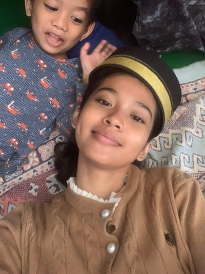Two individuals, a child and a young woman, are lying down in what appears to be a bedroom in Sungai Meriam, Indonesia. The child, positioned in the upper left of the frame, is wearing a blue pajama top with an orange crab print. The young woman, in the foreground, is wearing a brown cardigan with a white ruffled collar and a black hat with gold trim. The background shows bedding with a patterned design. The scene is a close-up selfie, suggesting the photo was taken by the young woman.