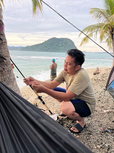 An overcast day at the beach in Babahie, Indonesia, features two men fishing. The foreground is dominated by a black hammock, partially obscuring a man squatting and attending to a fishing rod. He is wearing a light brown t-shirt and dark blue shorts. Next to him, another fishing rod is propped up on the pebbly shore. In the middle ground, a second man, wearing a blue cap and patterned shorts, stands closer to the water, also engaged in fishing. The ocean is a light turquoise, with waves gently lapping at the shore. In the background, a lush green island rises from the sea, its peak partially obscured by clouds. Palm trees with their fronds frame the top of the scene, hinting at a tropical setting.