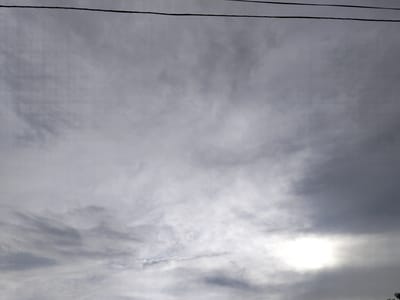 The image shows a cloudy sky above a faint silhouette of trees. A power line runs horizontally across the top of the frame. The clouds appear to be a mix of dark and light gray, with a bright patch of sunlight visible near the bottom right corner, suggesting it is daytime. The overall mood is somewhat somber due to the overcast conditions. The scene is likely outdoors, given the presence of the sky, clouds, and trees. No people or specific objects other than the power line are clearly discernible. There is no visible text in the image.