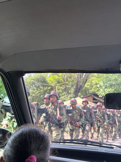 A group of soldiers wearing camouflage uniforms and helmets march in formation on a dirt road in Miruektaman, Indonesia. The soldiers are armed with rifles and batons. They are viewed from inside a vehicle, with the head and hair of a young person visible in the foreground. The background consists of trees and simple buildings.