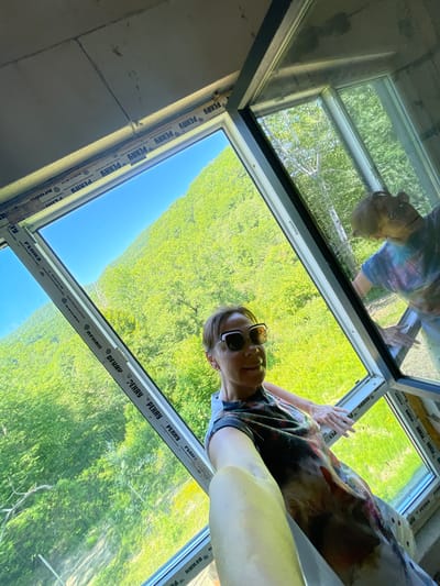 In Verkhniy Yurt, Russia, a woman is taking a selfie in a room with large windows offering a view of a lush, green hillside under a clear blue sky. She is wearing sunglasses and a colorful, abstract-patterned t-shirt. Her left hand is reaching out towards the window frame. The window frames have visible "PERRY" branding. Another person, partially visible in reflection on the window, is wearing a tie-dye shirt and appears to be cleaning the glass. The interior of the room is unfinished, with visible ceiling insulation and construction materials.
