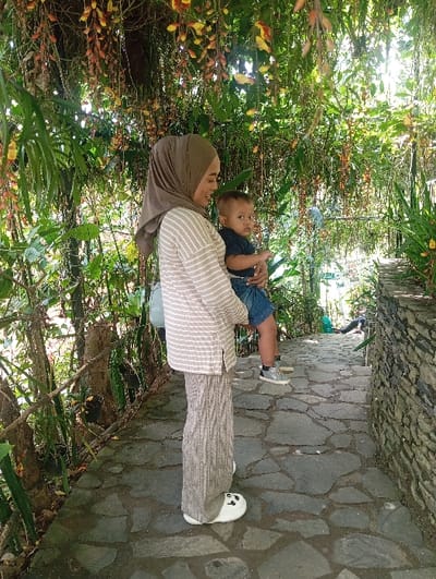 The scene depicts a female adult holding an infant on a stone pathway, stated to be located in Kajar, Indonesia. The female, appearing to be in her 20s or 30s, stands in profile facing right, her head turned slightly left, looking at the infant with a faint smile. She wears a plain, light brown hijab, a loose-fitting beige and white horizontally striped long-sleeved top, and loose-fitting, light brown or grey patterned trousers. Her feet are clad in white slip-on shoes featuring black panda-like designs. She supports the infant, approximately 1-2 years old, who faces directly towards the viewer, with his left arm around her waist and his right hand on her arm. The infant wears a dark blue t-shirt, blue denim shorts, and grey and black sneakers with white laces.

The pathway underfoot is constructed from irregular, flat grey flagstones, leading downwards and disappearing into the background on the right. To the right of the path, a low retaining wall made of stacked grey and brown stones is visible. To the left, a rustic fence made of rough wooden posts and rails runs parallel to the path, partially obscured by dense green foliage. The overhead canopy is lush with various green plants and numerous drooping vines bearing clusters of yellow and reddish-brown flowers. Sunlight filters through the dense foliage, creating dappled lighting on the subjects and the path.