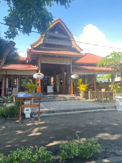 The image shows the exterior of a restaurant with a Balinese architectural style. The building features a steep, multi-tiered roof made of terracotta tiles, supported by wooden beams and columns. The facade is open, revealing seating areas with wooden furniture and bamboo blinds. Decorative elements include hanging lamps, umbrellas, and potted plants. A wooden stand displaying menus is positioned in the foreground, with a stone pathway and steps leading up to the entrance. The ground in front of the restaurant is covered with dark soil, and there are green bushes and trees scattered around the property. The sky is blue with a large, white cloud. The scene appears to be set in a tropical environment. This establishment is located in Buleleng, Indonesia.