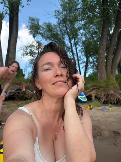 A medium shot captures a woman and a young man on a sandy beach near trees and a body of water. The woman is in the foreground, looking at the camera with a slight smile and her hand resting on her cheek. She has curly dark hair and is wearing a white bikini top and a gold necklace. Behind her, the young man is bent over, sticking his tongue out at the camera, with his bare chest and dark shorts visible. The background shows a grove of trees with exposed roots, a blue and green inflatable object, and scattered toy construction vehicles on the sand. The sky is partly cloudy. The presence of trees and sand suggests a natural outdoor setting, possibly a riverbank or lake shore. The location of Ol'khovskoye, Russia, is not visually indicated within the image itself.