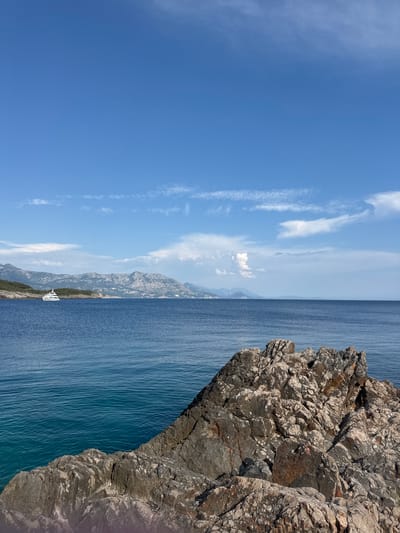 A sunny day unfolds over the coast of Dubraljevina, Montenegro, with a clear blue sky dotted with wispy white clouds. In the foreground, jagged brown rocks dominate the lower right quadrant, their textured surfaces catching the light. Beyond the rocks, the deep blue sea stretches out, meeting a distant, rugged coastline characterized by green hills and mountainous terrain. A white yacht is visible in the distance, anchored near the shore. The water is calm, reflecting the brilliant blue of the sky, and the overall scene conveys a sense of serene coastal beauty.