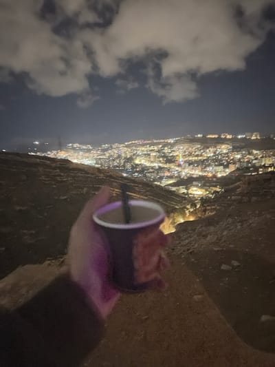 The image presents a nighttime scene overlooking a sprawling urban landscape identified as Dummar Municipality, Syria, from an elevated, rugged vantage point. In the immediate foreground, a human hand, illuminated by a distinct purple light source, holds a dark purple disposable cup containing a liquid and a black straw. The ground visible beneath the hand and extending into the midground consists of rough, unpaved, rocky terrain. Beyond this slope, the background reveals a vast array of artificial lights from buildings and infrastructure, indicating a city that extends across varied topography. The sky above is dark, with significant cloud cover that reflects ambient light from below.