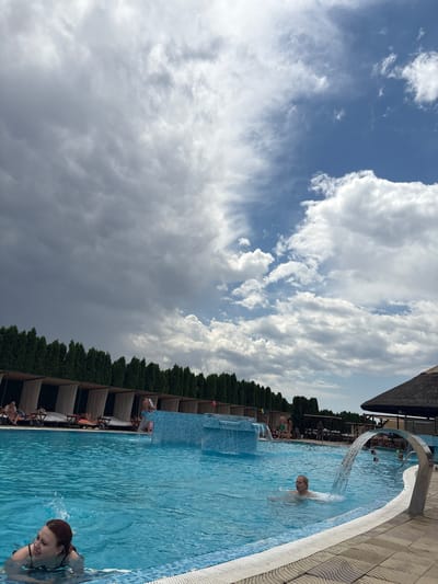 The image depicts a swimming pool area at a resort or water park. In the foreground, a woman with red hair is swimming in the clear blue water, looking to her left. Another person, a blonde individual, is visible further in the pool, near a waterfall feature. Several other people are scattered throughout the pool and in the background. The pool area features lounge chairs and cabanas, with a line of manicured evergreen trees forming a backdrop. A thatched-roof structure is visible to the right. The sky above is a mix of dramatic dark clouds and bright blue patches with white clouds. The ground around the pool is paved with patterned tiles. The scene suggests a leisure activity in an outdoor environment. This location is likely a resort or water park within the municipality of Stara Pazova, Serbia, given the provided context of its origin.