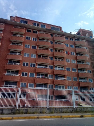 The image depicts a multi-story brick apartment building in Parroquia Guatire, Venezuela. The building has numerous balconies with white railings and white-framed windows. A white metal fence runs along the base of the building, separating it from the street. The sky is blue with some scattered clouds. The foreground includes a road and a curb with yellow paint. Clothing is hung to dry on some balconies. There are visible antennas on the roof of the building.
