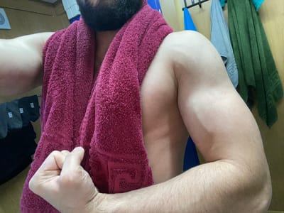 A man with a beard, wearing a maroon towel draped over his shoulder, flexes his bicep. He is in a room with a wooden wall. Several towels are hanging on a rack behind him, including a blue towel, a gray towel, and a green towel. A pair of black socks with white "NIKE" logos are visible on his left. The scene appears to be in a bathroom or dressing area.