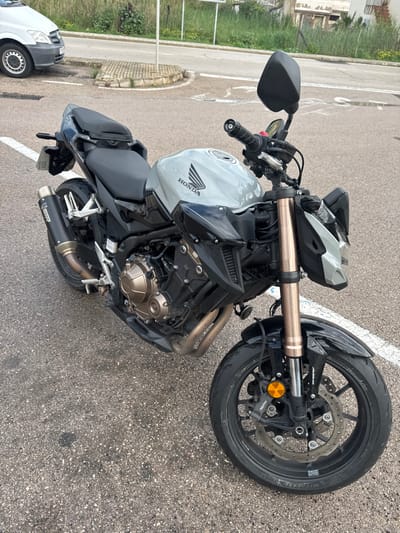 A light grey and black Honda naked motorcycle is positioned stationary on a damp asphalt surface, angled towards the front right, in Port D'andratx, Spain. The motorcycle features a light grey fuel tank with the "Honda" emblem, a black frame, black two-part seating, and a black aftermarket Akrapovič exhaust pipe on its left side. The engine casing and front suspension forks are finished in a bronze tone. Its black multi-spoke front wheel is equipped with a disc brake system and Nissin calipers. Water droplets are visible on the front fender and tire, suggesting wet conditions. White painted lines mark the asphalt parking area. In the background, a white Mercedes-Benz commercial van is partially visible parked to the left. A stone curb separates the pavement from a raised area with sparse vegetation, behind which a road and distant structures are discernible. No individuals are present in the scene.