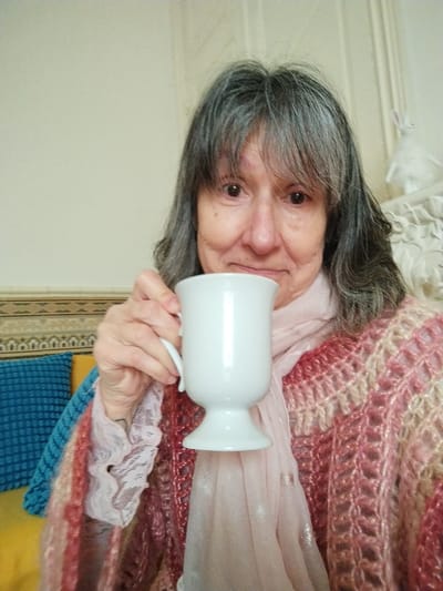 This image depicts an indoor scene located in Montélimar, France. The central subject is an adult female with graying brown hair, styled with bangs, looking directly at the camera with a faint smile. She is wearing a light pink, possibly lace, long-sleeved top, covered by an open-knit shawl in shades of red, pink, and cream, featuring shimmering threads. A sheer, light pink scarf is draped around her neck. Her right hand holds a white, footed ceramic mug with a handle, positioned in the lower center of the frame. In the background to the subject's left, a decorative patterned border is visible above a blue textured cushion and a yellow surface. To her right, a section of white, ornate carved architectural detailing is present, topped by a small white rabbit figurine. The wall behind the subject is a plain, light color.