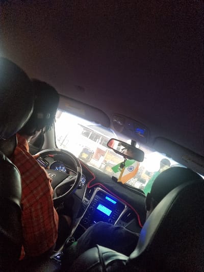 This image depicts the interior of a vehicle within Ojoto, Nigeria, viewed from a rear passenger seat looking forward. A male driver, wearing a red and white plaid shirt, is seated at the steering wheel, which bears a Hyundai emblem. His hands are positioned on the wheel. A second individual, a front passenger, is partially visible to the right, wearing a green shirt. The dashboard is accented with a red LED strip, and the center console displays an illuminated blue screen. An ornament resembling the Indian national flag hangs from the rearview mirror. Outside the windshield, a bright daytime urban environment is visible, featuring light-colored buildings and the presence of other individuals. The vehicle's interior seating visible in the foreground and passenger areas is black upholstered.