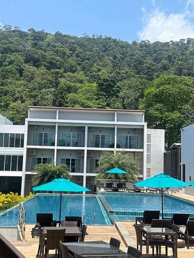 A horizontal eye-level shot reveals a multi-story resort building adjacent to a swimming pool, with a dense mountain range in the background. The resort building is white with a visible grid pattern. Each section of the grid contains windows. The swimming pool is long and narrow, running parallel to the building. Multiple teal-colored sun umbrellas are positioned next to the pool, alongside wicker-style tables and chairs. In the background, a steep mountain range is covered in dense green vegetation. The sky is light blue with a few white clouds. This scene is set in the city of Baan Klong Prao, Thailand.