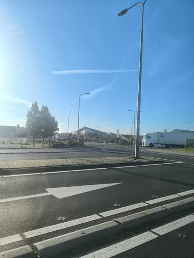 The image shows a bright, sunny day in Boskoop, Netherlands, with a clear blue sky and some wispy clouds. In the foreground, there is a road with a white arrow painted on it, indicating a direction. The road is bordered by a paved walkway and a concrete curb. In the background, there are several buildings, including a modern-looking structure with large glass windows. A white truck with a trailer is parked on the right side of the image. There are also trees and streetlights visible. The overall scene appears to be an industrial or commercial area.