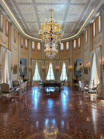 The image depicts a grand, ornate ballroom or reception hall. A large, multi-tiered crystal and gold chandelier hangs from the highly decorated ceiling, which features intricate plasterwork. The walls are painted a pale yellow and adorned with decorative moldings and large landscape paintings. Tall, narrow windows with diamond-shaped panes are evenly spaced along the walls, draped with heavy cream-colored curtains.
The room is furnished with an assortment of antique chairs and small tables, upholstered in floral and damask fabrics. In the center of the room, a long, dark-colored table with ornate legs is visible. The floor is made of polished dark wood with a repeating, intricate geometric and floral pattern, reflecting the light from the windows and chandelier. The overall impression is one of opulent historical grandeur. The location is the ballroom of the Mukhrani Palace in Mukhrani, Georgia. There are no people visible in the image.