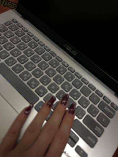 The image presents a close-up, slightly blurred view of a silver-gray ASUS laptop, featuring its keyboard and a person's hand. The laptop screen is dark, possibly off or displaying a black background. The keyboard is designed with light gray keys and dark gray inscriptions, clearly showing both English letters and Arabic characters, indicating a bilingual layout. The brand name "ASUS" is visible on the laptop's frame above the keyboard. A hand, adorned with long, dark red or burgundy manicured nails that have a faint lighter design, rests over the right side of the keyboard, suggesting the individual is either typing or about to type. The scene appears to be set indoors in Jaramana, Syria, consistent with the Arabic characters on the keyboard. The dim lighting implies the photo was taken during the evening or night.