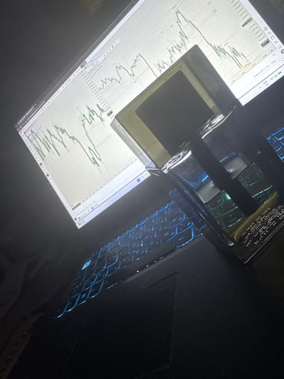 The image shows a monitor displaying a trading chart. A metal object, possibly an ashtray or a decorative piece, is placed on the monitor. A laptop keyboard with blue backlighting is visible to the right of the monitor. The image is dimly lit, and the angle suggests it was taken from above, looking down at the setup. No people or location details related to Morava, Bulgaria, are discernible in the image.