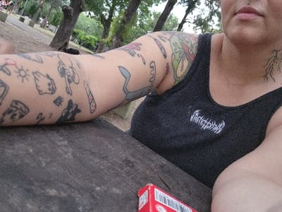 A person is captured from the shoulders up in a selfie, exhibiting extensive arm tattoos including a pizza slice, ghost, and sun. A large, colorful tattoo is prominent on the upper arm. The subject wears a black tank top with white lettering across the chest. A root-like tattoo is visible on the neck. A red pack with a visible barcode rests on a wooden surface. Trees and greenery are discernible in the blurred background, suggesting an outdoor setting. The person has a lip piercing.