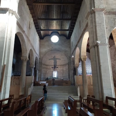 The image depicts the interior of the Cattedrale di San Ciriaco (Ancona Cathedral) in Ancona, Italy, viewed from the nave looking towards the apse. The scene is dominated by massive light-colored stone architecture, including round arches, thick columns with ornate capitals, and high walls. A wooden, coffered ceiling with exposed beams spans the nave.

In the foreground, rows of polished dark wooden pews line both sides of a central aisle, which has a light-colored, reflective floor. One individual, appearing to be a person with dark hair wearing a dark t-shirt and blue jeans, stands facing away from the camera on a wide set of stone steps leading up to the elevated altar platform.

The altar area features a simple wooden altar. Directly above and behind it, a dark crucifix with a visible corpus is suspended, positioned in front of a large, lighter cross silhouette integrated into the apse wall. A small rectangular window is visible below the suspended cross. Above this, a large circular oculus window is centered high in the apse, emitting bright light. Flanking the altar platform, large vertical organ pipes are visible within arched recesses on both the left and right sides of the nave. No other individuals are present. The overall environment is well-lit, suggesting daytime.