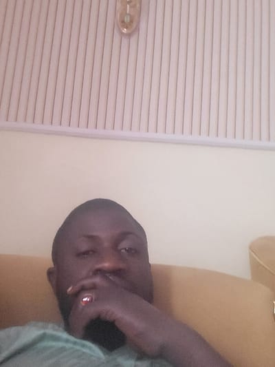 The image depicts a male individual indoors in Babawa, Nigeria. The subject, a dark-skinned male with short dark hair and a beard, is positioned in the lower-center of the frame, looking directly at the viewer. His left hand, adorned with a ring featuring a prominent reddish stone on the ring finger, is raised to his chin area. He is wearing a light green shirt. Behind and to his right, a portion of a mustard yellow upholstered armchair or sofa is visible. The background consists of a plain light beige wall in the mid-section, transitioning to a vertically grooved white or light-colored paneling in the upper section, which could be a ceiling or upper wall feature. A metallic, possibly brass-colored, elongated light fixture is mounted on this grooved paneling in the upper-right quadrant. The scene is illuminated by ambient indoor lighting.