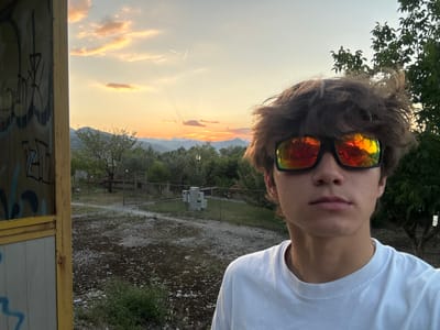 The image is a selfie taken outdoors, likely at dusk, with a young person wearing sunglasses and a white t-shirt in the foreground. The background features a rural or semi-rural landscape with trees, hills in the distance, and a dramatic sky with colorful clouds. To the left, a section of a structure with graffiti is visible, suggesting an urban or semi-urban setting. The overall impression is a casual, outdoor portrait set against a scenic backdrop. The location is Vranjina, Montenegro.