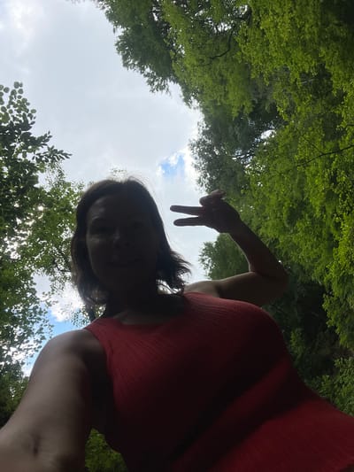 The media file is a low-angle, selfie-style shot looking up at a person and the sky, framed by lush green trees. The person, appearing to be female, is silhouetted against the bright sky and is wearing a red top. She is making a peace sign with her right hand, with two fingers extended. Her face is partially visible but shadowed. The scene suggests an outdoor setting, likely in a park or forest, with the sun shining through the leaves. The sky is partly cloudy, indicating a daytime setting. The overall mood seems relaxed and playful, possibly captured during a moment of leisure or enjoyment of nature. There is no visible text in the image. The location context provided is Khostinskiy, Russia.