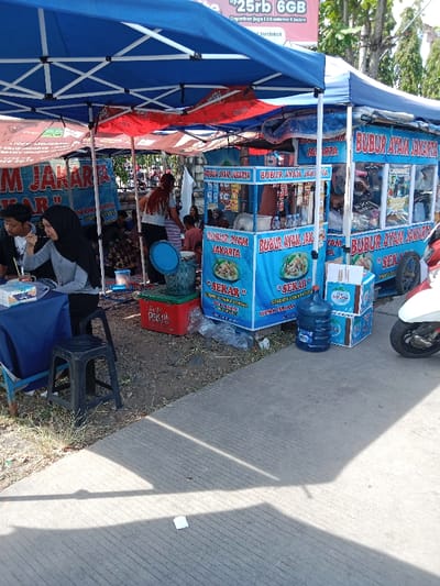 The scene is a street food vendor in Kendal, Indonesia, located at coordinates -6.931 latitude and 110.204 longitude. The vendor is selling "Bubur Ayam Jakarta", a type of chicken porridge. The vendor is situated under a blue awning, and there are other vendors nearby. There is a table with two women sitting at it. The women are wearing headscarves. One woman is holding a cup and drinking something. Behind the table are multiple blue and white signs with "Bubur Ayam Jakarta" written on them, as well as other signs advertising food. There are also several blue and white coolers with drinks in them. On the right side of the image, there is a white motorbike parked. There is a red cooler in the background. The ground is a light grey concrete. There is a white napkin on the ground in the foreground. The environment is typical for an outdoor food vendor in a city.