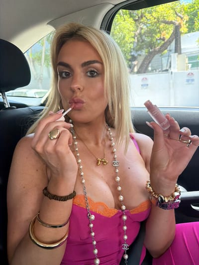 A woman with blonde hair is applying lip gloss while inside a vehicle, likely a car. She is wearing a pink top with orange lace trim and several pieces of jewelry, including necklaces, bracelets, and rings. The lip gloss appears to be a Kylie Cosmetics product. Outside the car window, trees and a building are visible. This scene is located in West Hollywood, California, United States.