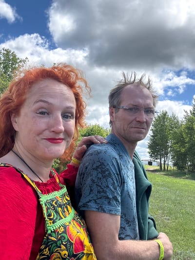 In a selfie taken outdoors, a woman with bright red, curly hair and prominent freckles smiles at the camera. She wears a red top and a colorful apron with a floral and folk-art pattern. A man with tousled hair and glasses stands to her right, looking into the distance. He is wearing a blue t-shirt with a black abstract pattern and has a green jacket draped over his shoulder. Both individuals appear to be outdoors in a grassy area with trees in the background, under a partly cloudy sky. The scene is likely the city of Tri Ozera, Russia, suggested by the overall environment and cultural attire of the woman.
