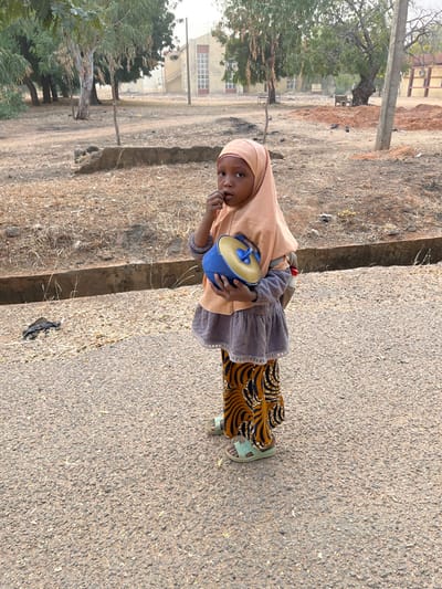 A young girl in a peach hijab, a grey dress with ruffled edges, and patterned pants stands on a paved surface. She holds a blue and yellow container with handles. She gazes at the camera while putting a finger in her mouth. The background consists of dry vegetation, a concrete ditch, and a building with white-framed windows in the distance. A partially visible tree stands to the left of the frame.