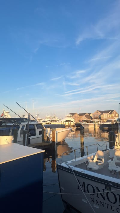 This is a vertical, eye-level, outdoor photograph of boats docked in a harbor in Nantucket, Massachusetts, United States of America, under a clear blue sky with thin, wispy clouds. In the foreground, the stern of a white boat with the name "MON" partially visible in large black lettering, followed by "NANTUCK" in smaller letters, is moored to a dock. The boat has white seats and stainless-steel railings. To the left of this boat is a blue-and-white structure.
Behind this foreground boat, other boats are docked in the harbor. Some have fishing poles attached. In the middle ground, a row of buildings with brown shingled roofs and wooden siding lines the waterfront. These buildings appear to be shops or residences. The water in the harbor reflects the sky and the boats. The lighting suggests it is either early morning or late afternoon, with the sun casting a warm glow on the scene.