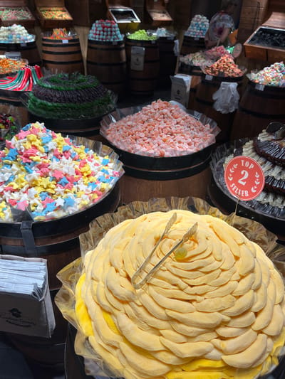 The image depicts a candy store display. Candies are arranged in barrels, presumably for sale. The candies include yellow banana candies, small star-shaped candies, orange candies, grape-shaped candies and small flower-shaped candies. A sign reads "Top 2 Seller" is visible. A pair of tongs are on top of a pile of yellow banana candies. Napkins branded "Captain Candy" are also present. The scene is likely in a commercial area, such as a candy store, potentially in Verona, Italy, based on the prevalence of candy stores in tourist locations.