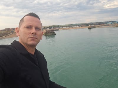 A man is taking a selfie in front of a body of water. He is wearing a black textured jacket over a black t-shirt. The man is facing the camera, and his expression is neutral. Behind him, there is a body of water that appears to be the sea. In the background, there is a coastline with buildings, a beach, and some structures in the water that look like a pier or a breakwater. The sky is overcast with clouds. The scene appears to be a daytime shot, likely late morning or afternoon. The location is suggested to be Adur, United Kingdom. The overall mood of the image is calm and perhaps a bit contemplative. There is no visible text in the image.