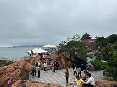 The image captures a coastal scenic area within Xiaoyushan Park, Shinan District, Qingdao, China, under an overcast, light-grey sky.
In the foreground, reddish-brown rock formations dominate the lower left and center, interspersed with a light-colored, square-tiled pedestrian pathway. Approximately ten individuals, primarily adults and children of East Asian ethnicity, are present here; some walk along the path, others stand or sit on the rocks. On the far left, a young girl in a pink top is visible. A group of three individuals, including a child in a yellow top, sits on a rock formation on the right side of the path.
The midground features more reddish-brown rocks leading down to a calm, blue-grey sea. A metal railing and a narrow walkway extend along the coastline. On a platform near the water, three white, conical-roofed tents are erected, beneath and around which multiple individuals are gathered. Further to the right, green trees and shrubs are interspersed with natural and possibly artificial rock structures, including a dark, cave-like formation.
The background consists of the sea on the left, with two light-colored vessels visible in the distance and a faint outline of land on the horizon. Above the midground vegetation and rocks, a prominent multi-tiered, traditional Chinese-style pavilion with a green roof and dark eaves, identified as the Wangchao Pavilion (Wave-Listening Pavilion), is situated atop a tree-covered hill. A smaller, red-domed structure is visible to the right of the pavilion. The scene is populated by numerous visitors engaged in leisure activities such as walking, observing, and gathering.