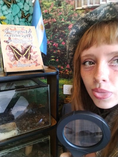 A woman in a hat sticks her tongue out, holding a magnifying glass. Beside her is a book with the title "Mariposas Argentinas" and an image of a butterfly. An Argentinian flag is in the background. A small terrarium sits on a shelf. The backdrop features a nature scene with flowers and butterflies.