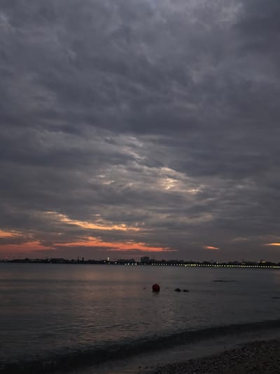 This is a vertical, low-angle, full shot of a seascape at dusk. The sky is filled with dark, dramatic clouds with hints of orange and yellow from the setting sun peeking through. The water is dark and slightly choppy, reflecting the moody sky. In the distance, a city skyline is visible, illuminated by numerous lights that form a continuous line along the horizon. A bright red buoy is partially submerged in the water, near two dark, indistinct shapes that could be rocks or small boats. The beach in the foreground is dark and appears to be made of pebbles. The overall mood is peaceful and serene, with a touch of mystery due to the dim light and overcast sky. There is no visible text.