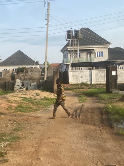 A man in a striped t-shirt and dark pants walks across a dirt road in Iddo Tudunwada, Nigeria. He is the central figure in the image, walking from left to right, with his body angled towards the viewer. In the background, two modern houses are visible. The house on the left has a dark, pitched roof and appears to be under construction, with stacks of concrete blocks in front of it. The house on the right is a multi-story building with a dark roof, balconies, and water tanks on a metal structure attached to its side. Utility poles with overhead wires are present in the sky, indicating the presence of electricity infrastructure. The ground is a mix of dirt, sand, and sparse green vegetation. The scene suggests an ongoing construction or development area within a residential neighborhood.