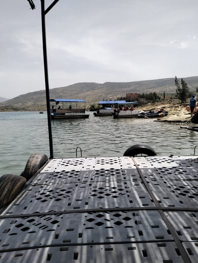 The image depicts a tranquil scene at Bin El Ouidane, Morocco, featuring a lake with several boats. A floating platform constructed of gray plastic modules secured with metal bands is in the foreground, with tires attached to the side acting as bumpers. In the water, two covered pontoon boats are visible, with several people visible on one of the boats. A small motorboat is docked near the shoreline, where people are also gathered. The background reveals a hilly landscape with sparse vegetation and buildings. The sky is overcast, suggesting a muted, diffused light. The overall impression is a tourist or recreational area on a relatively calm day.