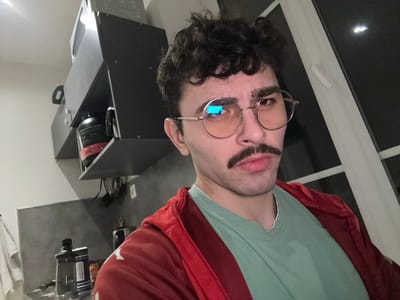 A person with curly brown hair, a mustache, and round, light-tinted glasses looks at the camera. They wear a light green t-shirt and a red hoodie. Behind them are dark kitchen cabinets with items on the shelves, including a black canister with red accents. A white window with a dark background is visible on the right side. The overall lighting is somewhat dim.