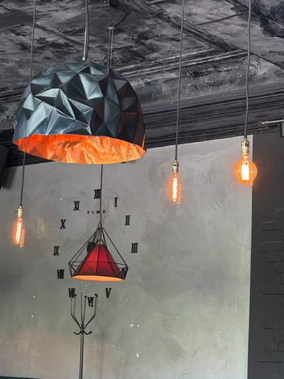 This media file displays a dimly lit interior with a focus on decorative lighting and an abstract art piece. The scene appears to be a commercial space, possibly a restaurant or bar, with a modern industrial aesthetic.

The primary objects are several Edison-style light bulbs, some round and glowing warmly, others elongated and emitting a similar orange hue. These bulbs are suspended by wires, contributing to the industrial feel. A large, faceted, geometric object hangs in the foreground, its exterior a dark, angular material, while its interior is lined with a shimmering, textured orange material. This object resembles a deconstructed lantern or a sculptural element.

In the background, a large clock face is subtly integrated into the grey wall. Roman numerals mark the hours, though some are partially obscured or difficult to discern. The wall itself has a textured, concrete-like finish, with a darker, more distressed section on the left side of the frame, possibly a textured wall covering or painted effect. The overall lighting is low, creating a moody and atmospheric environment. There are no people visible. The time of day and weather are not discernible from the image. No text is clearly visible besides the Roman numerals on the clock. The location context of Yelizovo, Russia, does not offer any specific visual cues within the image itself.