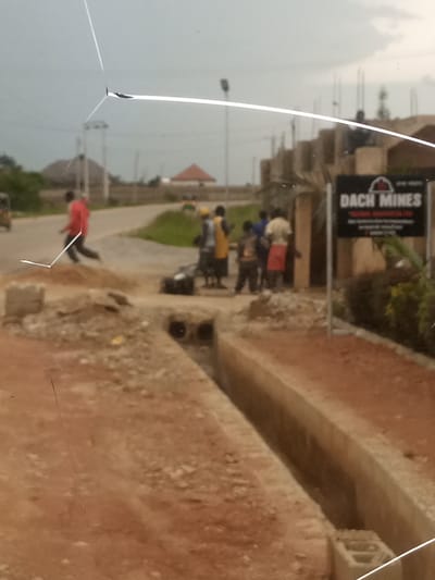 The image captures a street scene in Reifild-Ist, Nigeria, likely during the daytime under an overcast sky. A group of people are visible, some appearing to be workers. One individual in a red shirt is running, while others stand in small clusters. There are several buildings under construction in the background, indicated by exposed structural elements and scaffolding. A sign with the text "DACH MINES" and "NATURAL RESOURCES LTD" is prominently displayed on the right side of the frame, suggesting a business or mining operation is nearby.
The foreground shows a partially constructed drainage ditch or trench, with exposed soil and concrete elements. A small, dark object, possibly a piece of machinery or equipment, is situated near the ditch. The overall atmosphere appears to be one of ongoing development or work. The presence of multiple people in the frame suggests a community or work environment. The sky is a muted gray, indicating cloudy weather, but there are no immediate signs of rain. The road in the middle ground shows some traffic, including a yellow vehicle and possibly a tricycle. The scene is characterized by a blend of construction, daily activity, and commercial signage, providing a glimpse into the local environment of Reifild-Ist.
