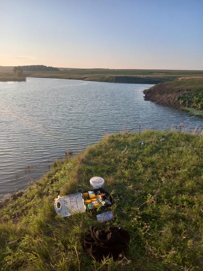 The image shows a tranquil natural scene on the outskirts of Polevye Ineli, Russia, beside a calm body of water. The sun is low in the sky, casting a warm, golden light across the landscape, creating long shadows and highlighting the texture of the grass and water. On the grassy bank of the water, fishing equipment is laid out, including two fishing rods propped up by orange holders, a tackle box filled with various fishing lures and accessories, and a white bucket. A dark fabric, possibly a jacket or blanket, is resting on the grass near the tackle box. The water is a deep blue, with gentle ripples on its surface, reflecting the clear, pale blue sky. The opposite bank of the water is lined with green vegetation and more distant fields, suggesting a rural or semi-rural setting. The overall impression is one of peaceful solitude, likely a scene of a fishing trip or a moment of quiet contemplation in nature.