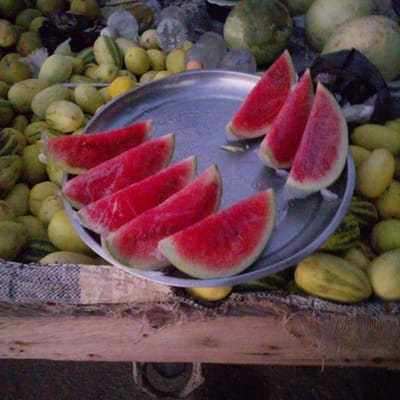 This image depicts a close-up view of a metal platter filled with sliced watermelons, displayed outdoors, possibly at a market stall in Gulak, Nigeria. The platter is filled with several triangular watermelon slices, arranged in a fanned pattern. The vibrant red flesh of the watermelon is clearly visible, with some white rind remaining. In the background, a variety of other fruits, including what appear to be more watermelons and possibly other melon varieties, are piled up. The fruits are a mix of green and yellow colors, with some exhibiting striped patterns. The overall setting suggests a vendor display of fresh produce. The lighting is natural, indicating an outdoor scene. No people are directly visible in the frame, but the arrangement of the fruit implies human activity for sale.