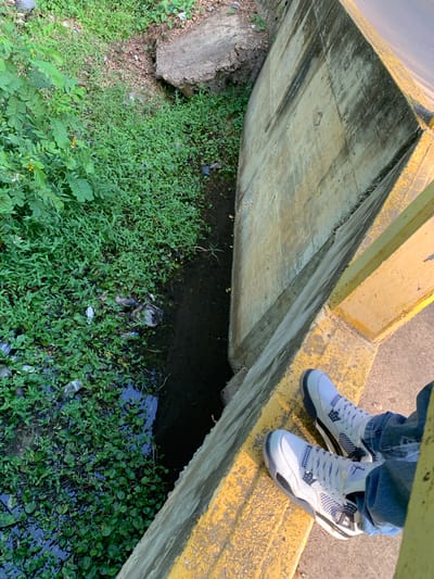 A high-angle, medium shot captures a person's feet and legs clad in blue jeans and white sneakers, standing on a yellow-painted concrete bridge railing. The perspective looks down into a narrow, overgrown drainage channel. Lush green vegetation, including small round leaves and larger leafy plants, fills the channel, which also contains dark water and visible trash, such as plastic bottles and bags. A large, rough concrete structure juts into the upper left of the frame, partially overgrown with vegetation. The concrete wall of the channel is stained and weathered, with visible signs of age and water damage. The scene appears to be an outdoor urban or semi-urban environment, possibly in El Valle del Espíritu Santo, Venezuela, given the context. The lighting suggests it is daytime, with sunlight casting shadows.