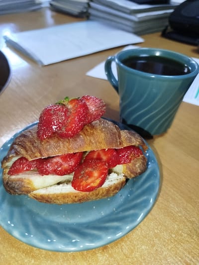 A close-up, eye-level, indoor shot captures a croissant sandwich filled with strawberries and cream, presented on a teal plate, resting on a light brown wooden desk. To the right of the sandwich, a teal mug filled with dark coffee is visible. In the background, stacks of papers and office supplies are blurred. The scene is set in an office environment, likely within the city of Navapolatsk, Belarus.