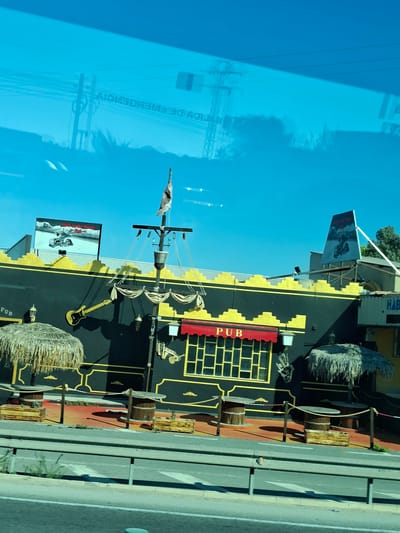 The image shows a building designed to resemble a pirate ship, located in the city of San Fulgencio, Spain. The ship-like structure is black with yellow accents and features a mast with a flag, a ship's wheel, and decorative ropes and pulleys. There are several tables and chairs outside the structure, some under thatched umbrellas. A sign above the entrance reads "PUB." In the background, there are power lines and a clear blue sky. The reflection of power lines and signage is visible on the blue tinted glass covering the building.