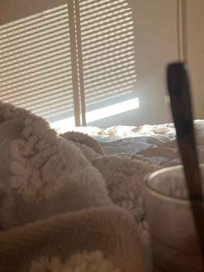 An indoor scene in Hardthausen am Kocher, Germany, depicts a bed and a wall illuminated by natural light. In the foreground, a textured, light-colored, possibly cream or off-white, fluffy blanket occupies the lower left and central portions of the frame. To the far right foreground, a blurry, dark, slender object extends vertically, partially obscuring a blurry, light-colored, cylindrical container, likely a glass or cup. The mid-ground reveals crumpled, light-colored bedding, consisting of sheets and a duvet. The background features a plain, light-colored wall with prominent horizontal shadows cast upon it by sunlight entering through a window equipped with venetian blinds. Two vertical lines within the shadow pattern indicate window frame elements. No individuals are present. The primary action involves sunlight casting distinct patterns onto the interior surfaces.